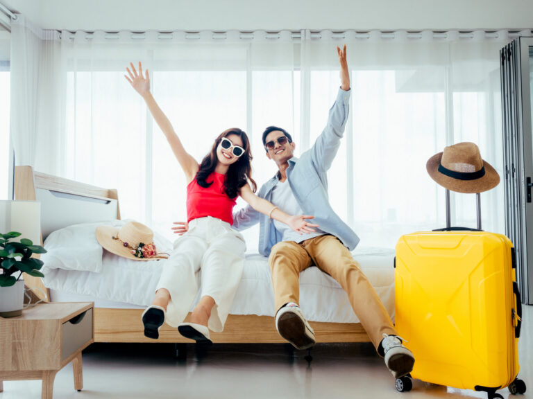 Ready To Travel, Happy Holiday, Check In. Summer Vacation Concept. Asian Couple Raising Hands With Joy, Man And Woman Wear Sunglasses Smile With Happy On White Bed With Yellow Suitcases On A Trip.