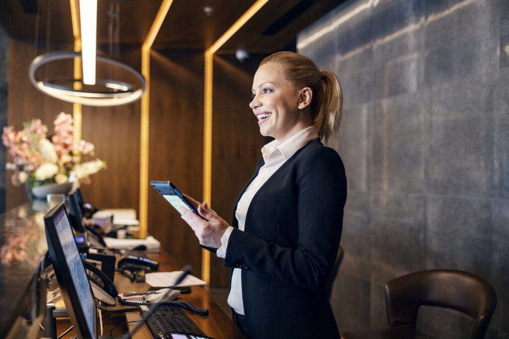 A happy receptionist is talking with hotel guest and making a reservation on a tablet.